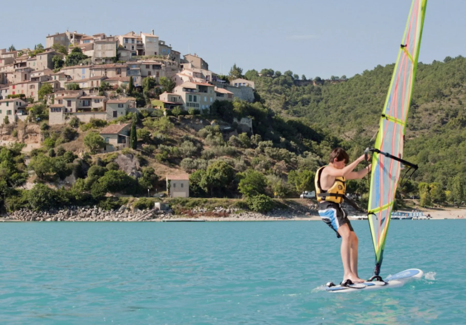 Planche à voile sur le lac de Castillon devant un village perché